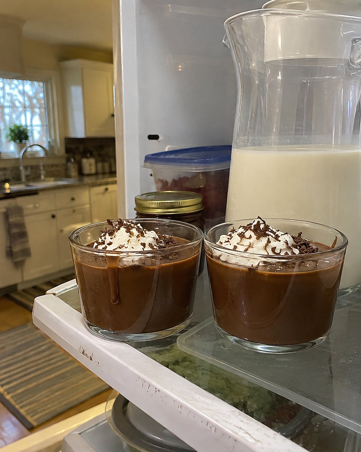 Two homemade chocolate pudding cups on a refrigerator shelf beside a pitcher of milk.