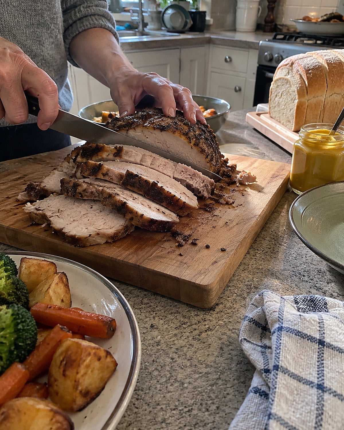 A sliced Sunday roast on a board beside a loaf of sandwich bread and a jar of mustard.