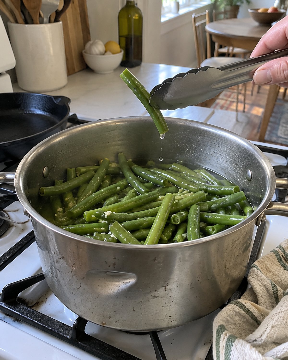 A dented pot of green beans on a white stove, with tongs holding one bean above the rim.