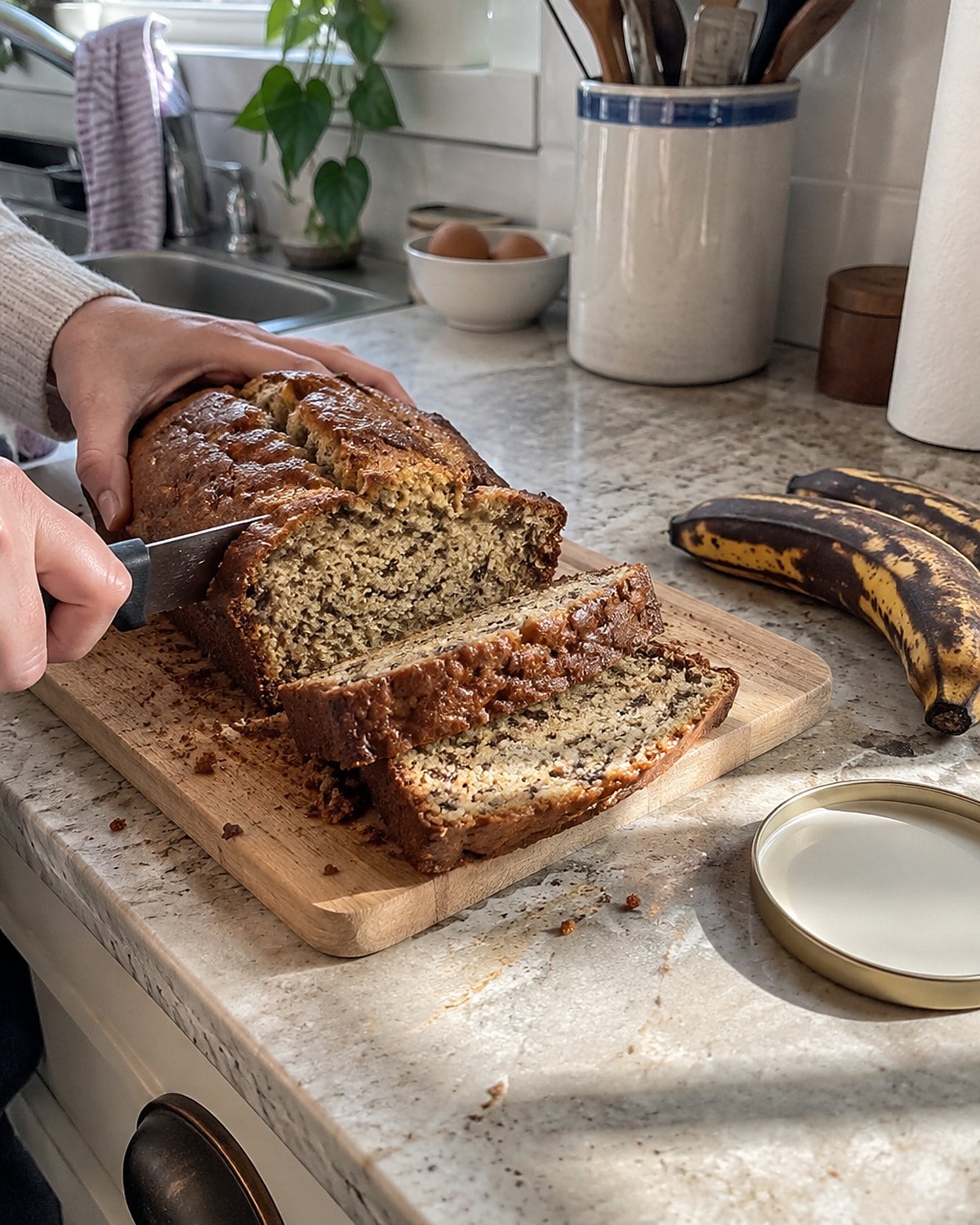 A partly sliced loaf of banana bread on a counter beside two dark bananas.