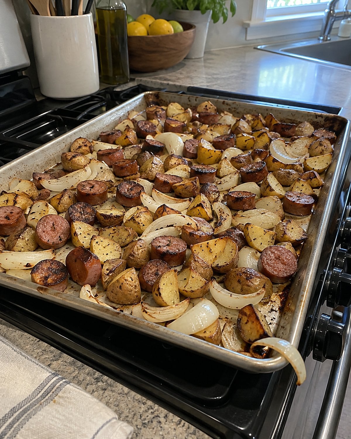 A sheet pan of sausage, potatoes, and onions on a stovetop, with browned pieces left unevenly spaced.