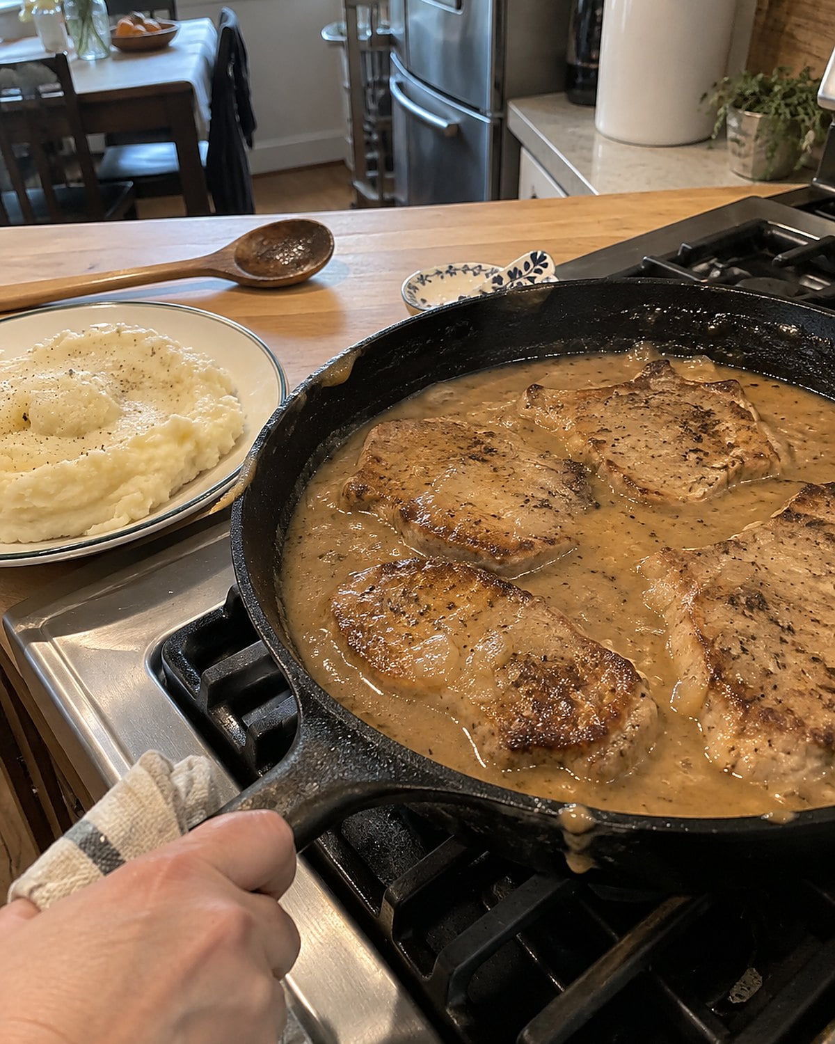 Pork chops in gravy in a skillet, with mashed potatoes waiting on a plate nearby.