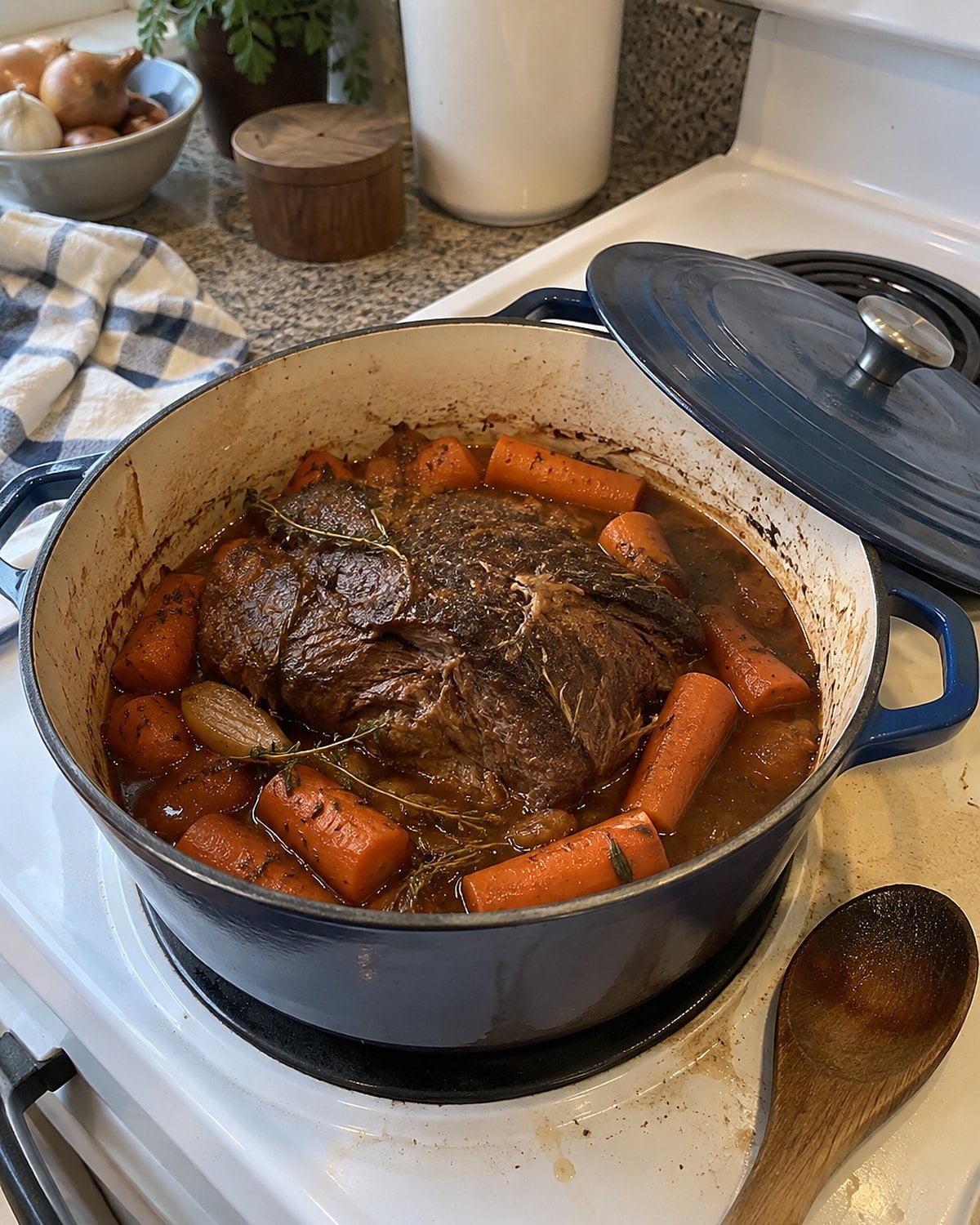 A Dutch oven of pot roast and carrots on a white stove, with the lid set crooked beside it.