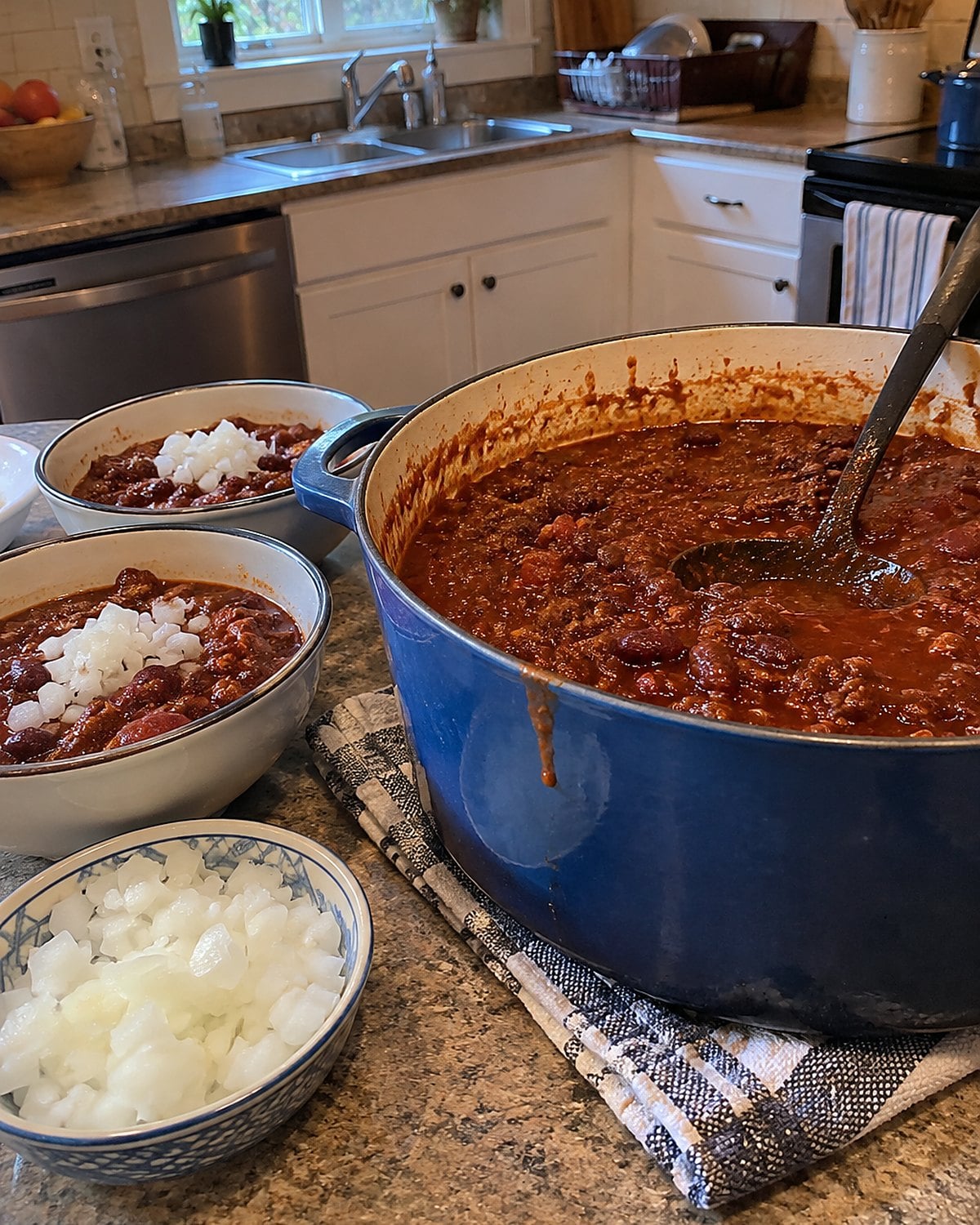 A pot of chili beside bowls and a small dish of chopped onion on a kitchen counter.