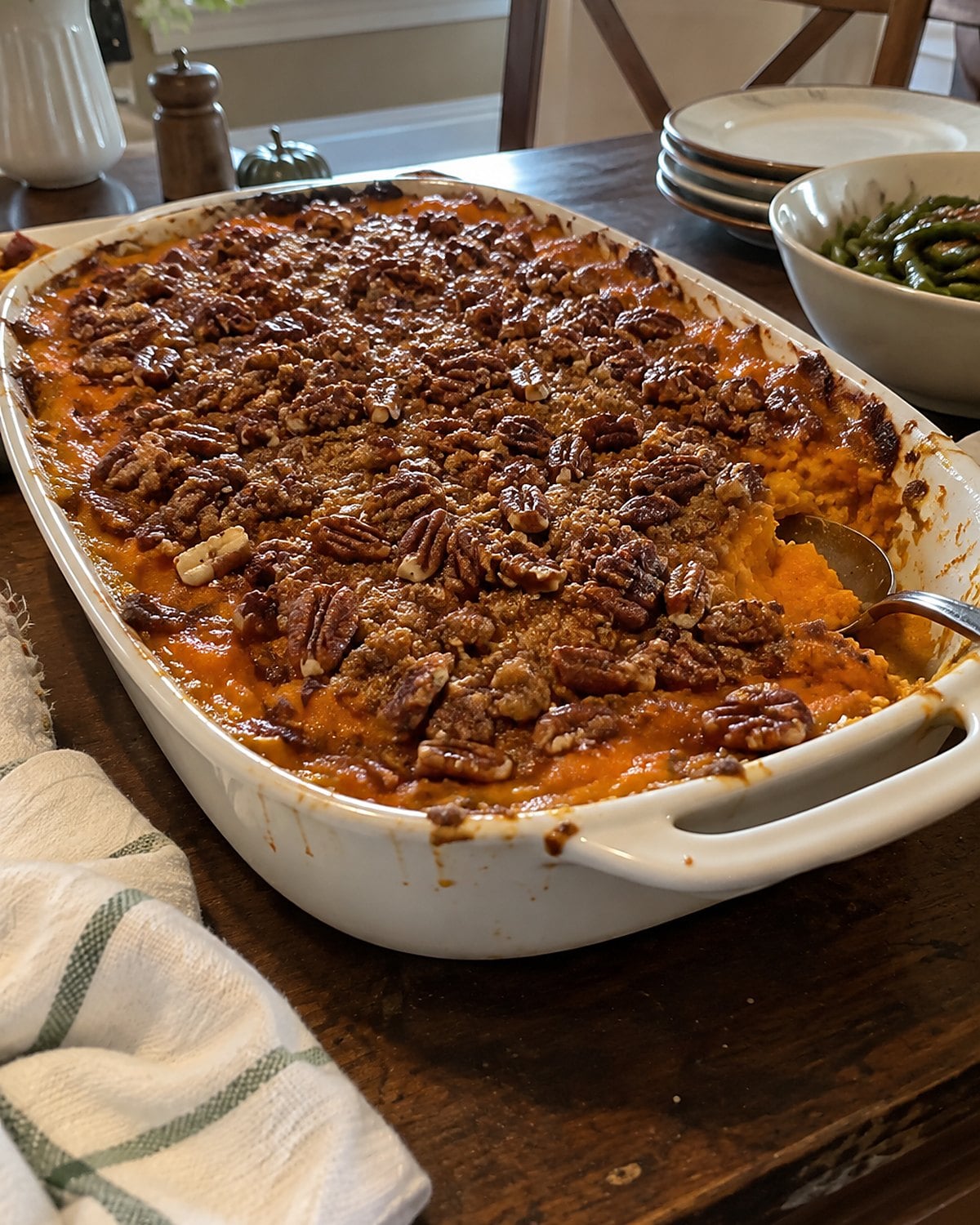A sweet potato casserole with a pecan topping on a holiday sideboard, with a serving spoon tucked beneath its edge.