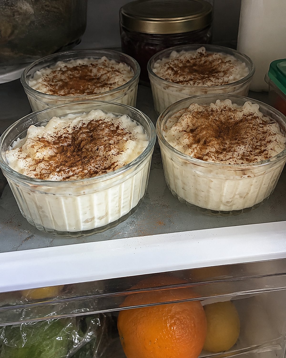 Custard cups of rice pudding on a fridge shelf, with cinnamon scattered unevenly over them.