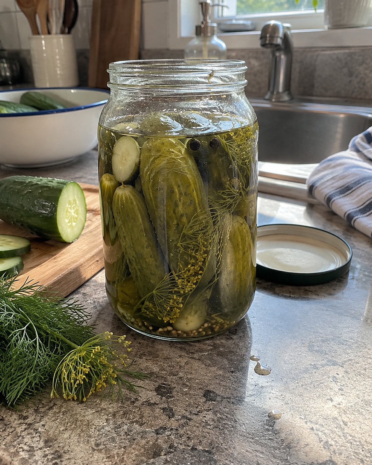 An open jar of dill pickles at the sink, with a cut cucumber and sprig of dill nearby.
