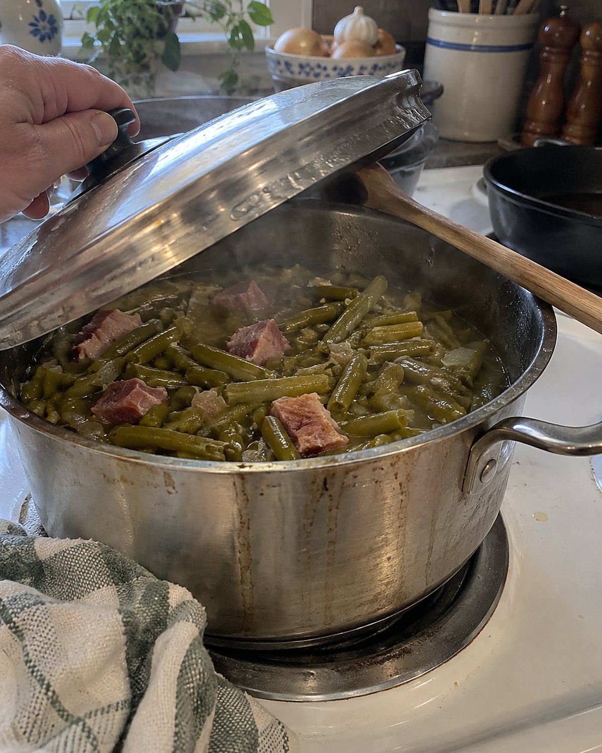 A dented pot of green beans and ham on a white stove, its lid propped on a spoon.