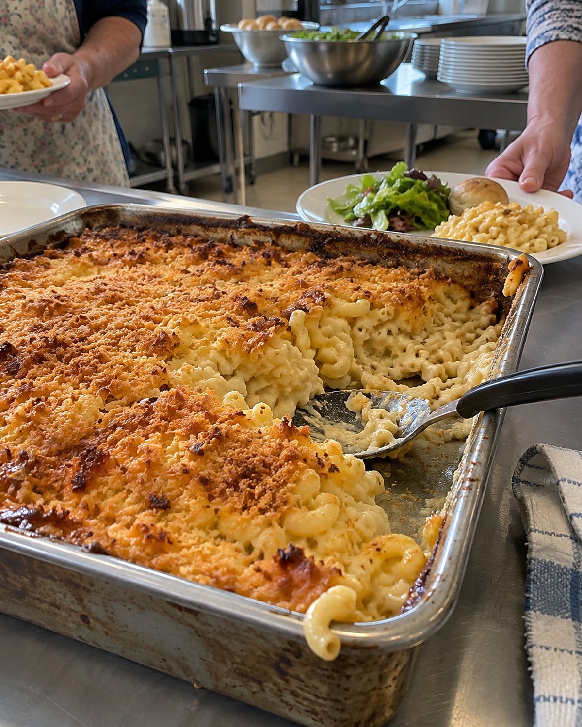 A partly served macaroni casserole on a church hall table, with a folded dish towel beside its pan.