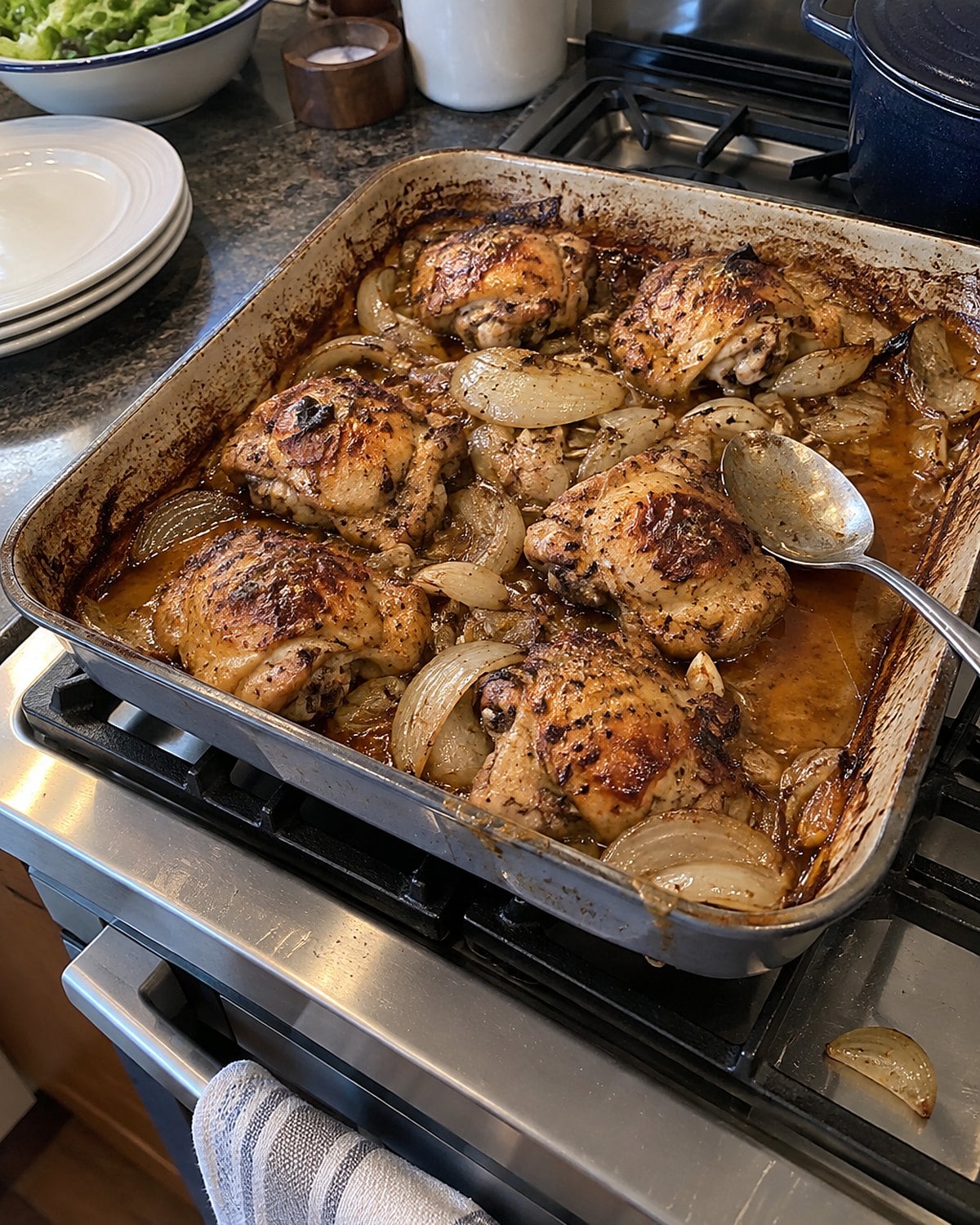 A roasting pan with chicken and browned onions on the stove, a spoon resting in the pan juices.