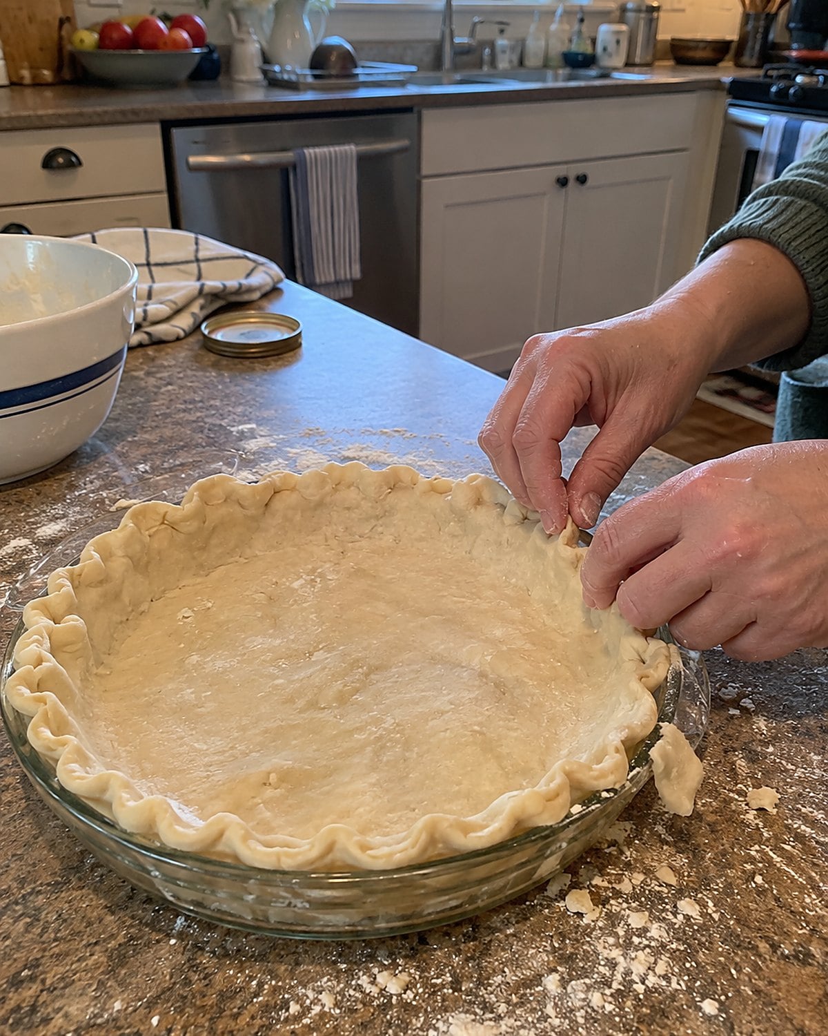 A pie crust with an uneven crimp in a glass plate, with flour scattered across the nearby counter.