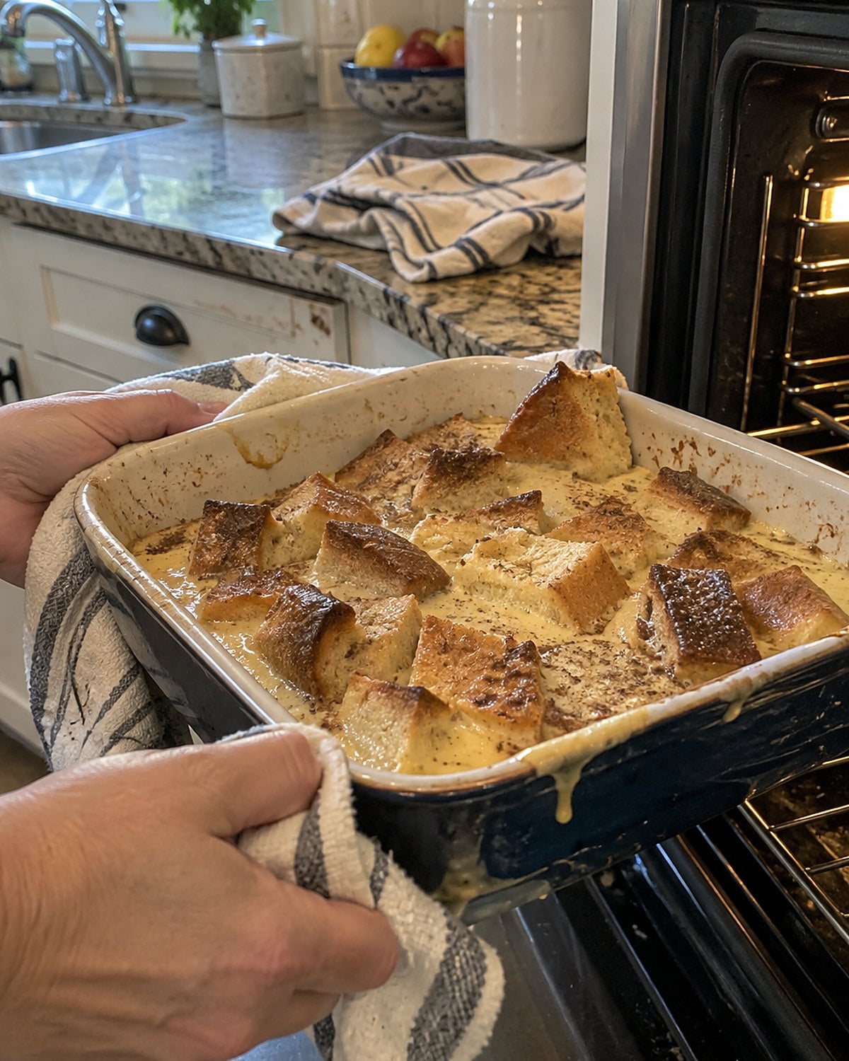 Torn bread ends soaking in custard in a worn baking dish beside an open oven.