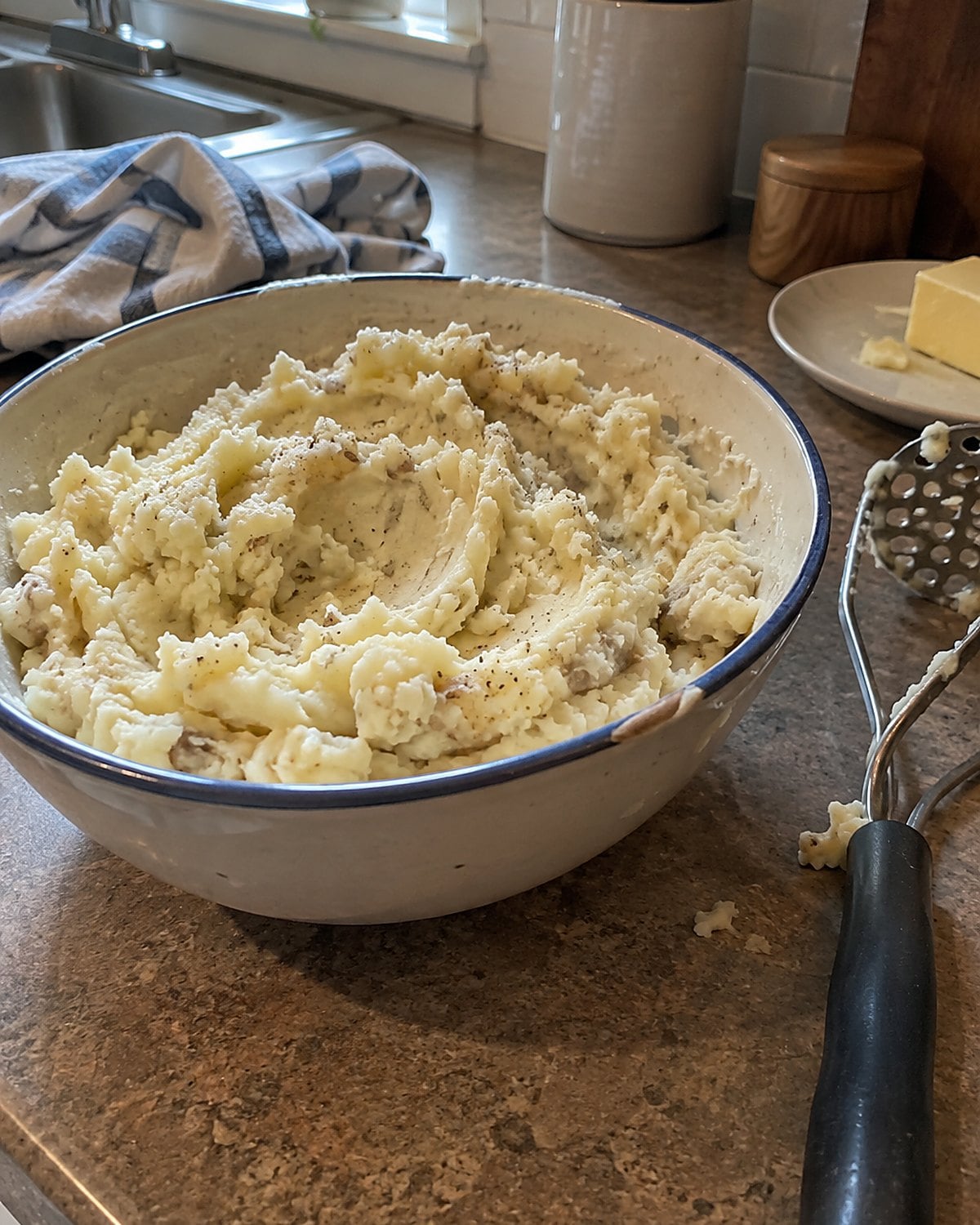 A chipped serving bowl of mashed potatoes with small lumps and a hand masher resting beside it on the counter.