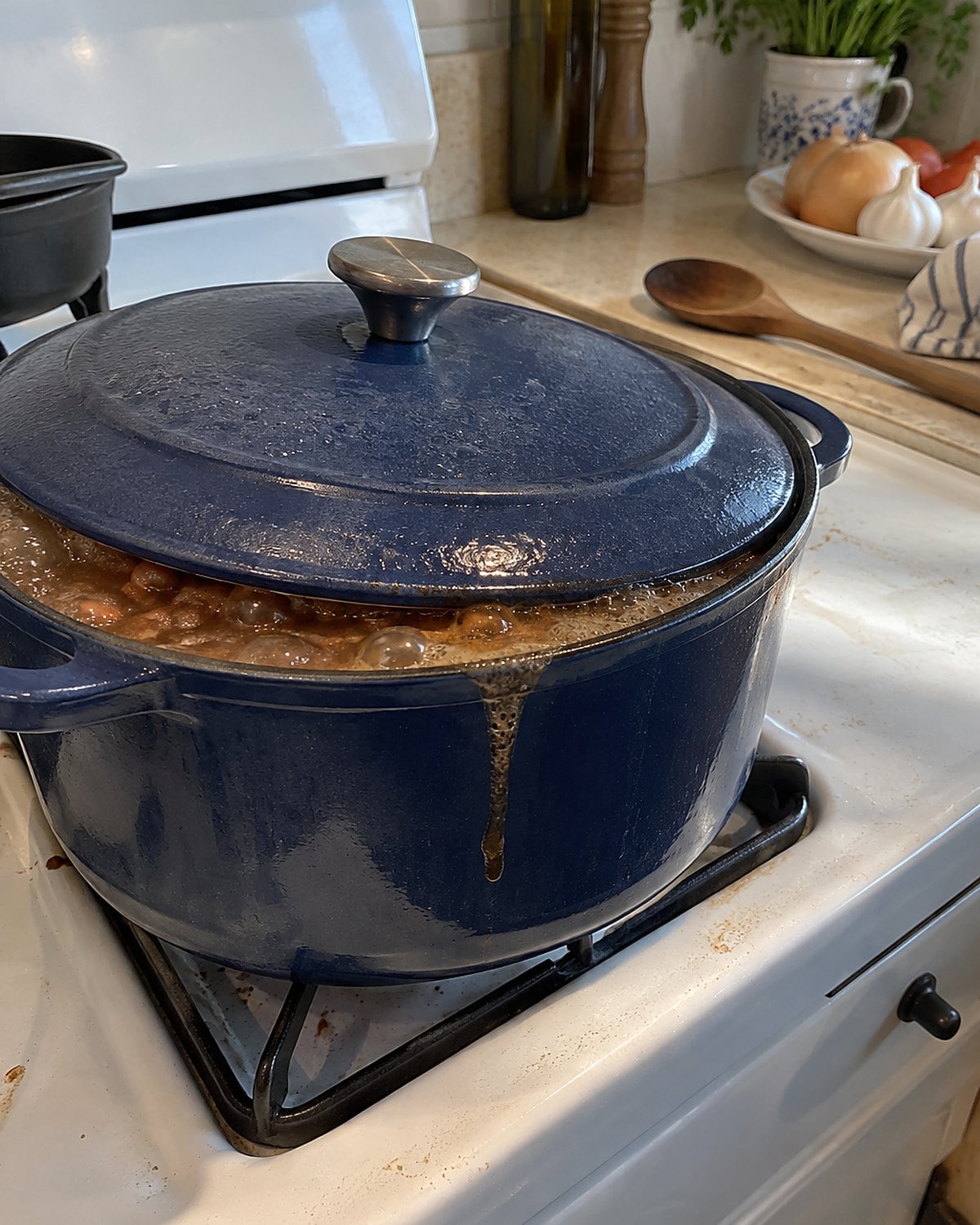 A lidded pot of beans on a white stove, with broth bubbling around the edge of the slightly crooked lid.