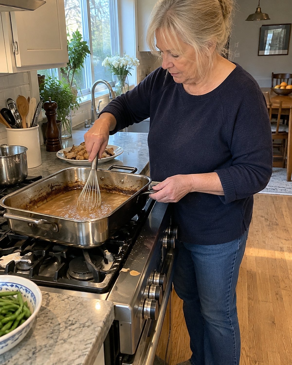 A hand whisking gravy in a roasting pan on the stove, with browned drippings along the rim.