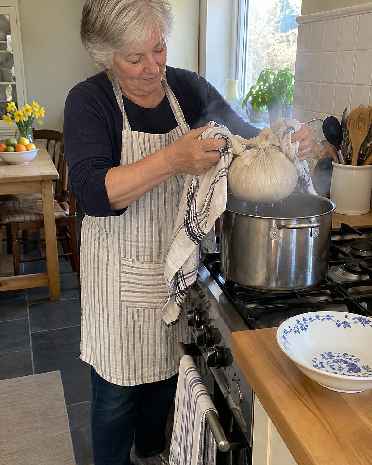 A cloth-covered pudding mold being lifted from a steaming pot with thick kitchen towels.