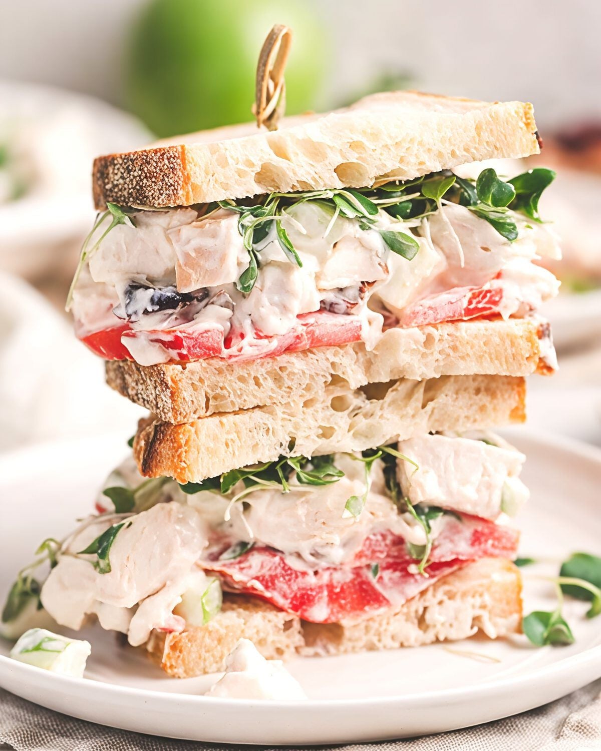 A tall chicken salad sandwich cut in half and stacked high on a white plate, layered with creamy chicken salad, fresh sliced tomatoes, and microgreens between crusty artisan bread.