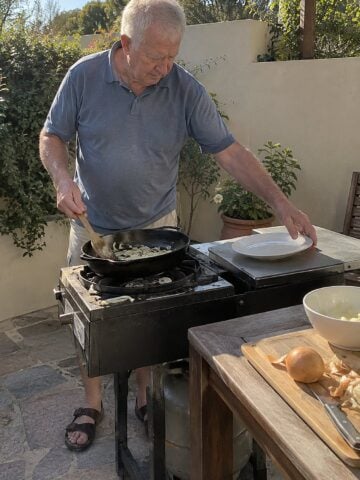 A man in his seventies turns food in a cast-iron pan on an outdoor burner in mid-afternoon, with his other hand reaching for a plate beside the stove.