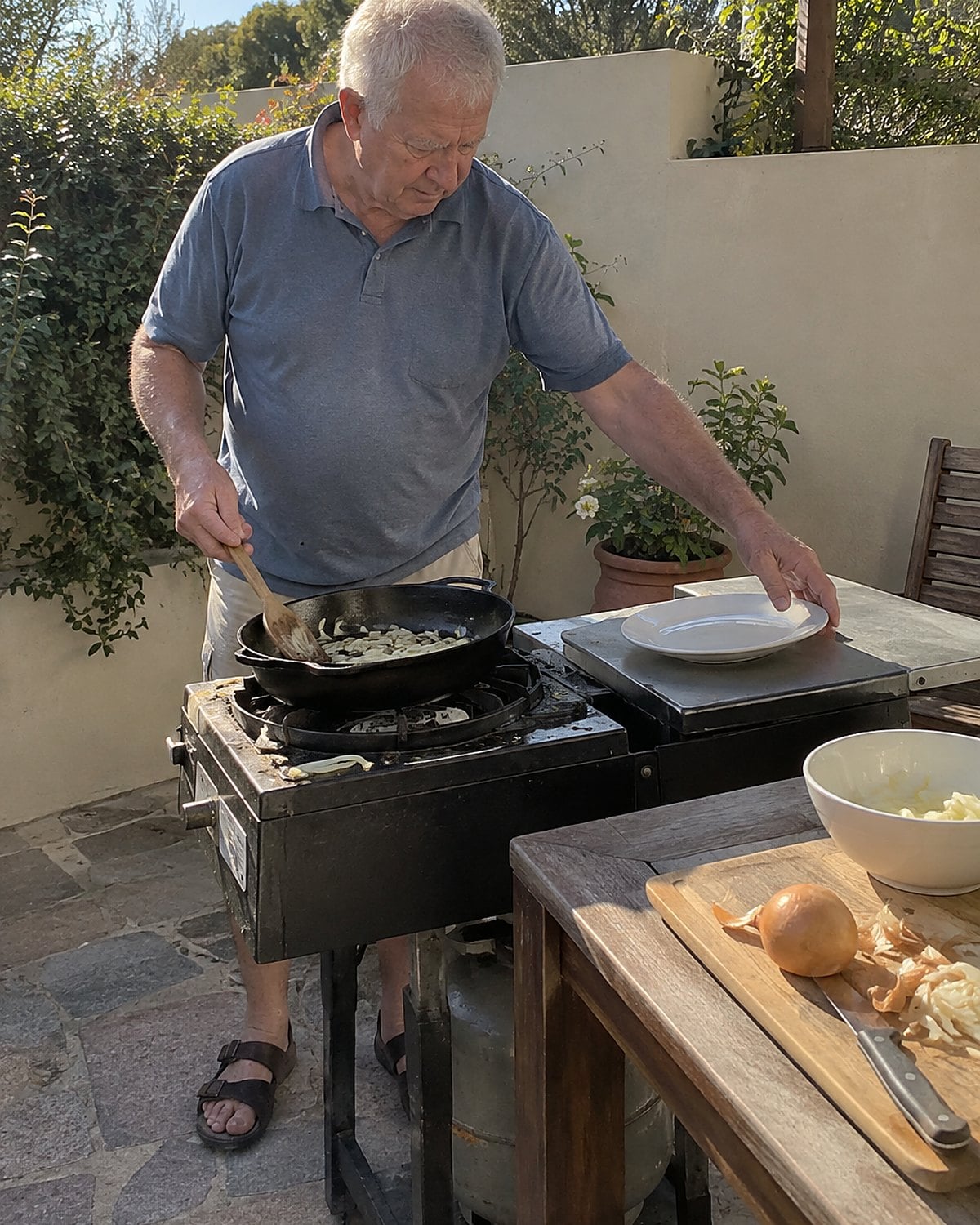 A man in his seventies turns food in a cast-iron pan on an outdoor burner in mid-afternoon, with his other hand reaching for a plate beside the stove.