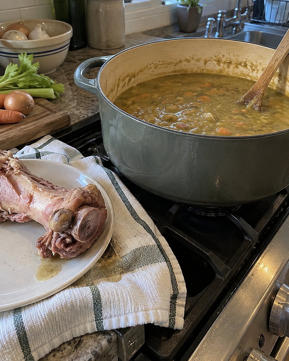 A ham bone rests beside a pot of thick split pea soup on the stove.