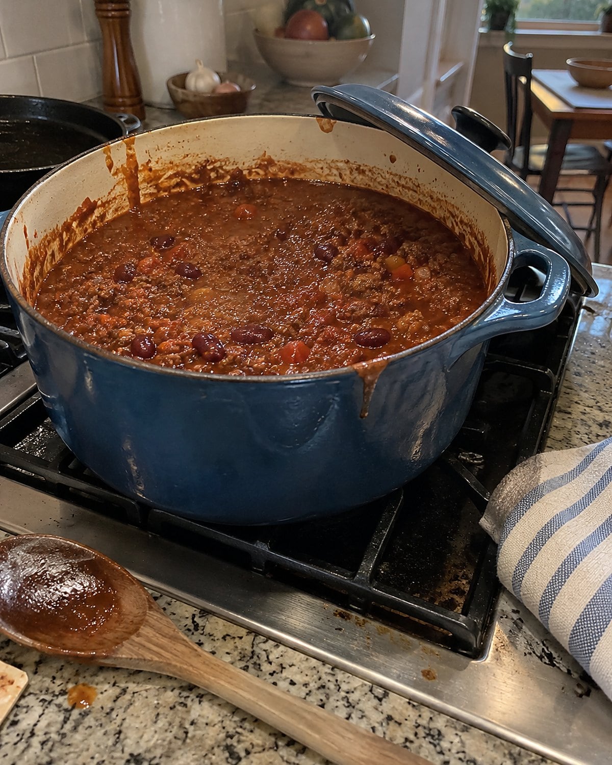 A dented Dutch oven of chili sits on the stove with its lid tipped against the handle.