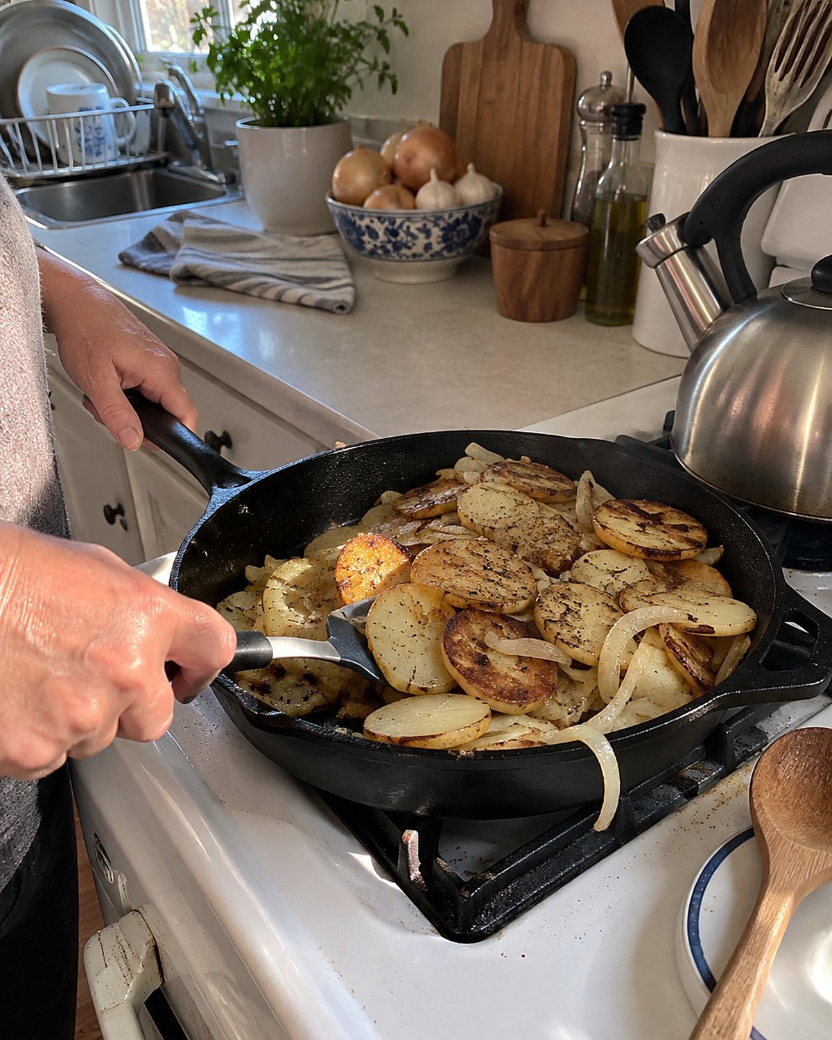 Sliced potatoes and onions crowd a dark skillet on a white stove, with a spatula tucked beneath one edge.