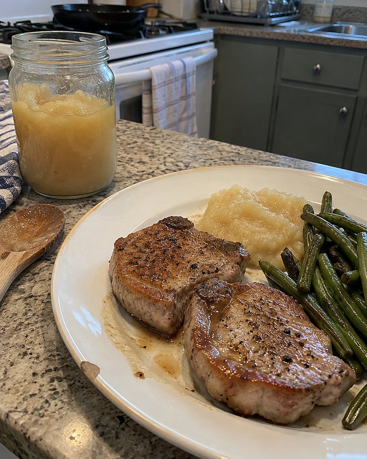 Pork chops rest on a plate beside an opened jar of applesauce.