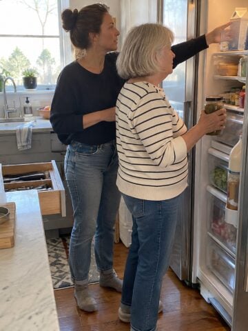 At the open fridge door in mid-afternoon, a woman holds a jar while her adult daughter reaches past her for a carton, neither looking at the camera.