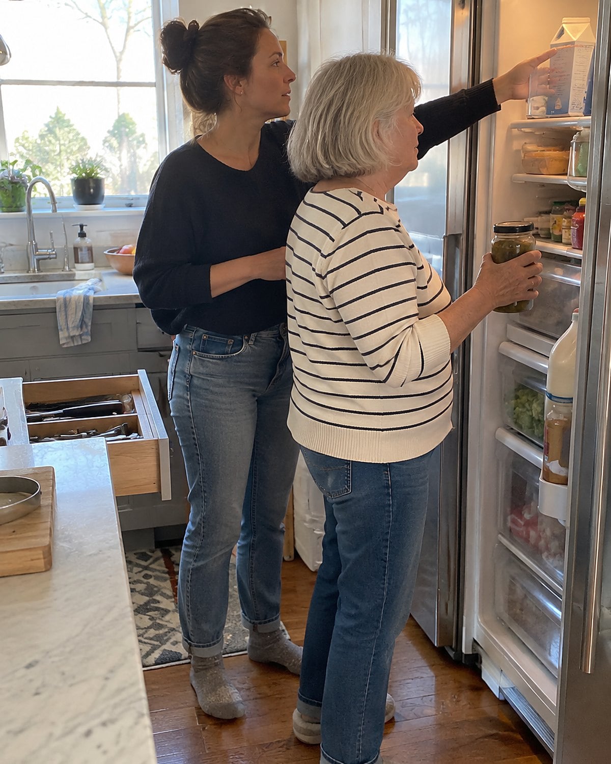 At the open fridge door in mid-afternoon, a woman holds a jar while her adult daughter reaches past her for a carton, neither looking at the camera.