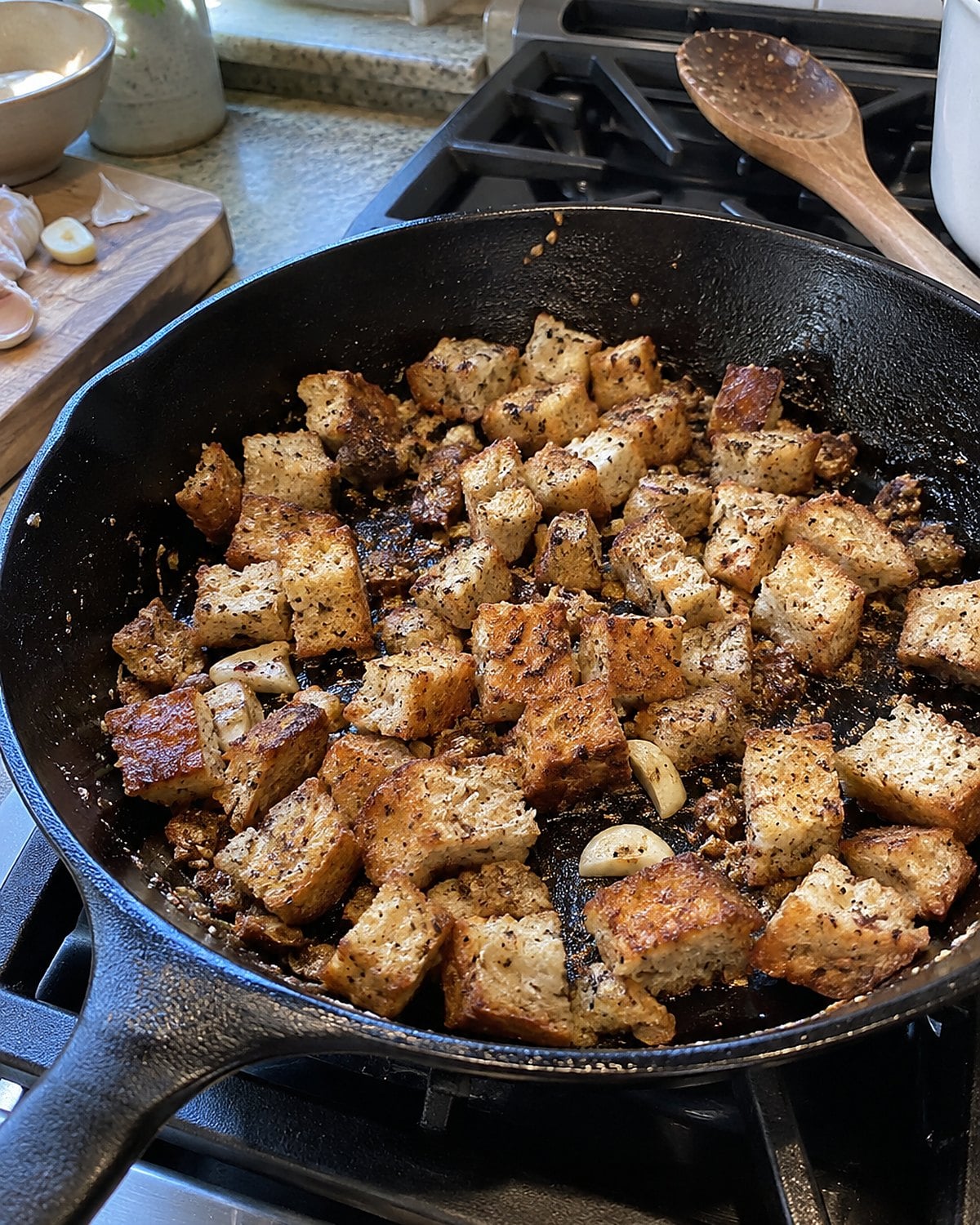 Bread cubes browning in a skillet beside a cut garlic clove.