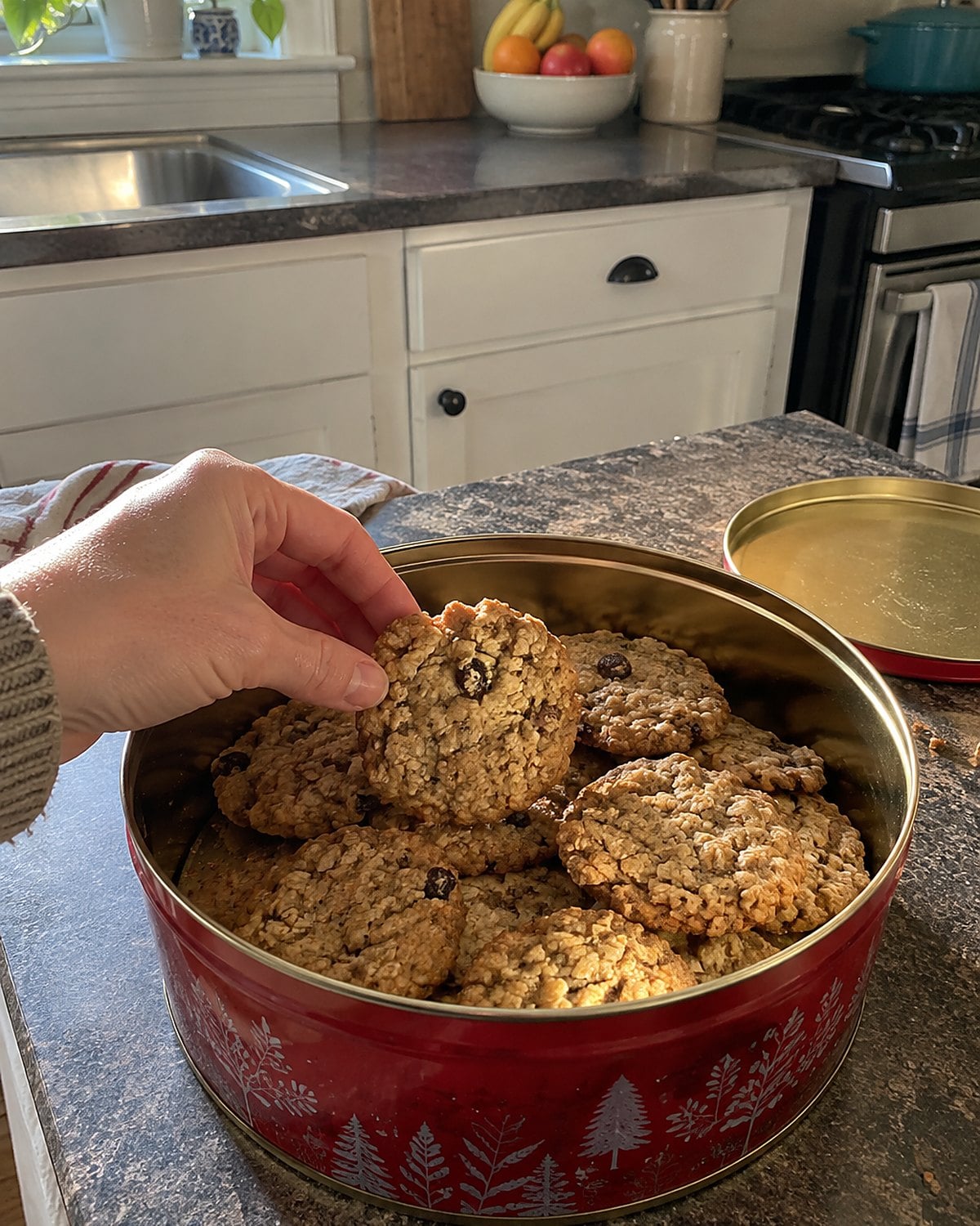 An open cookie tin with uneven oatmeal cookies on a kitchen counter.