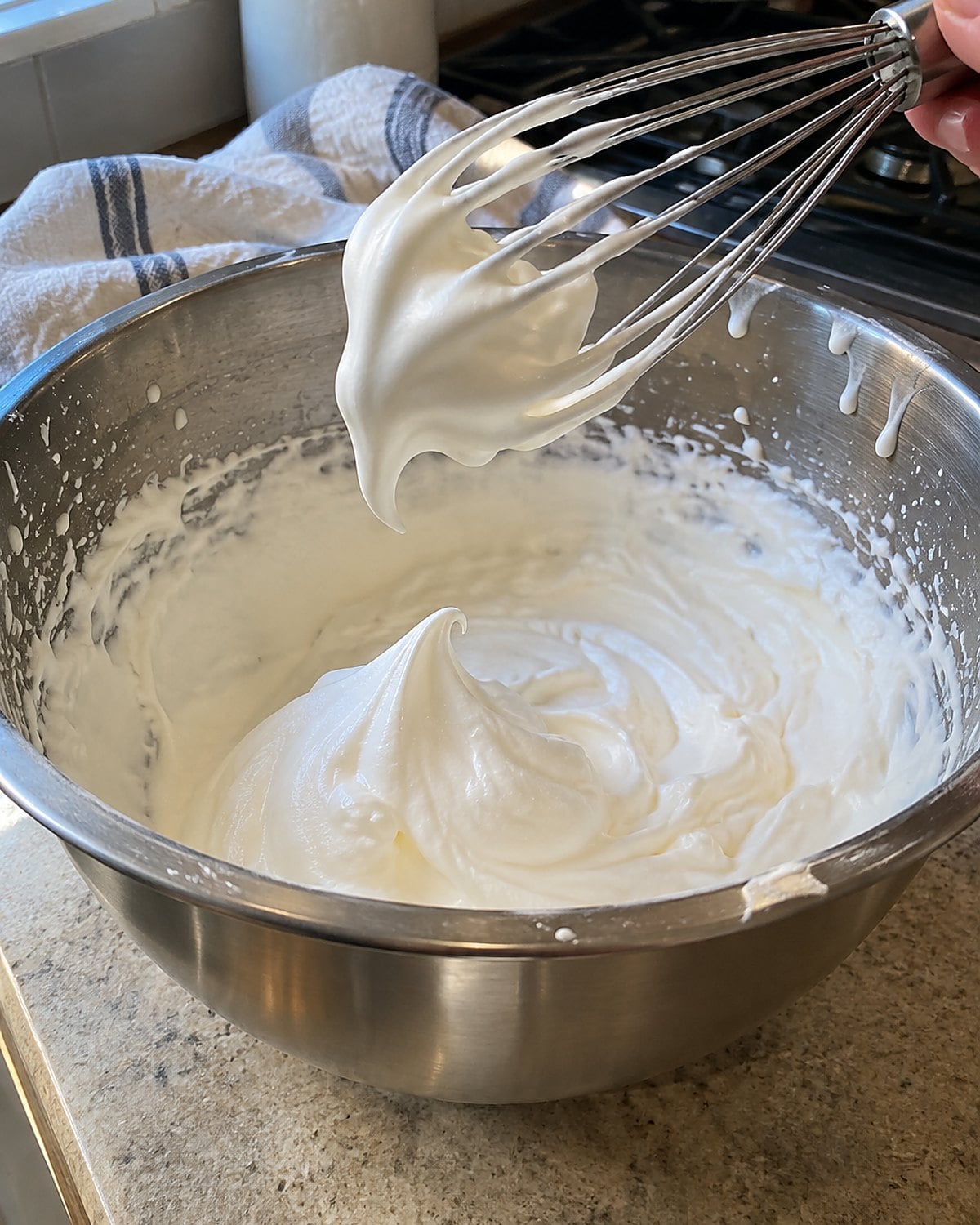 A whisk lifted from a metal bowl of cream, with one bent peak visible.