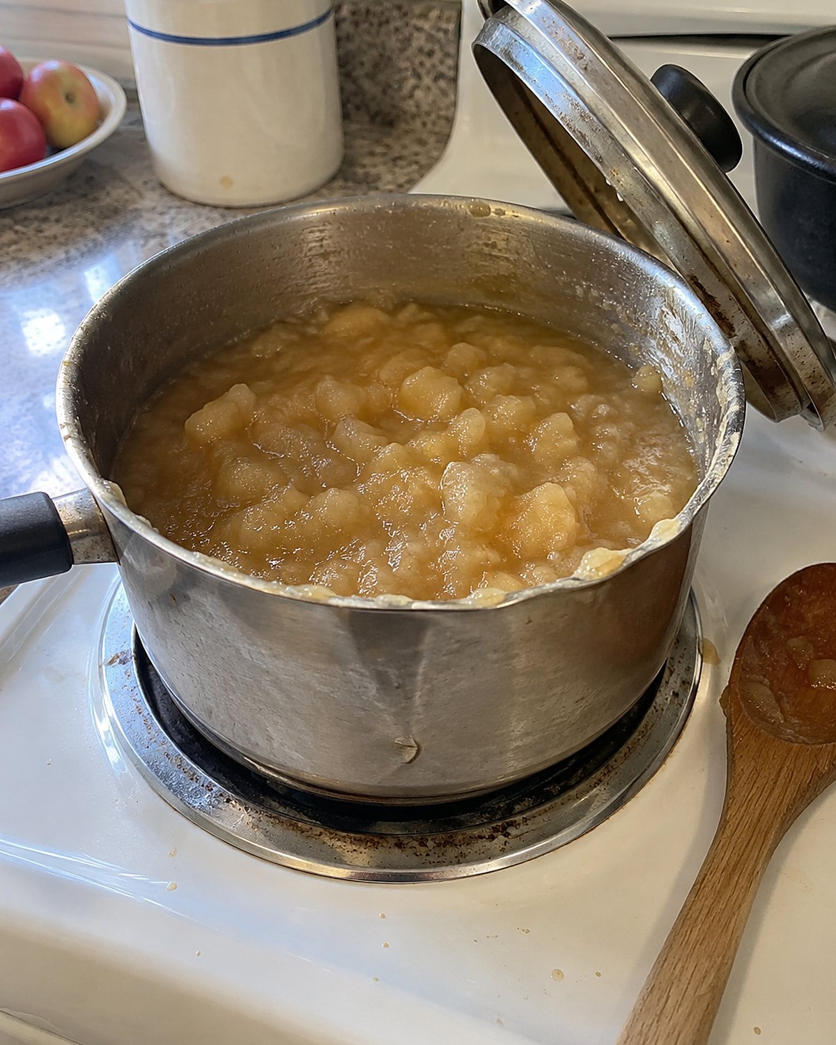 A dented saucepan of chunky applesauce on a white stove, its lid propped open.