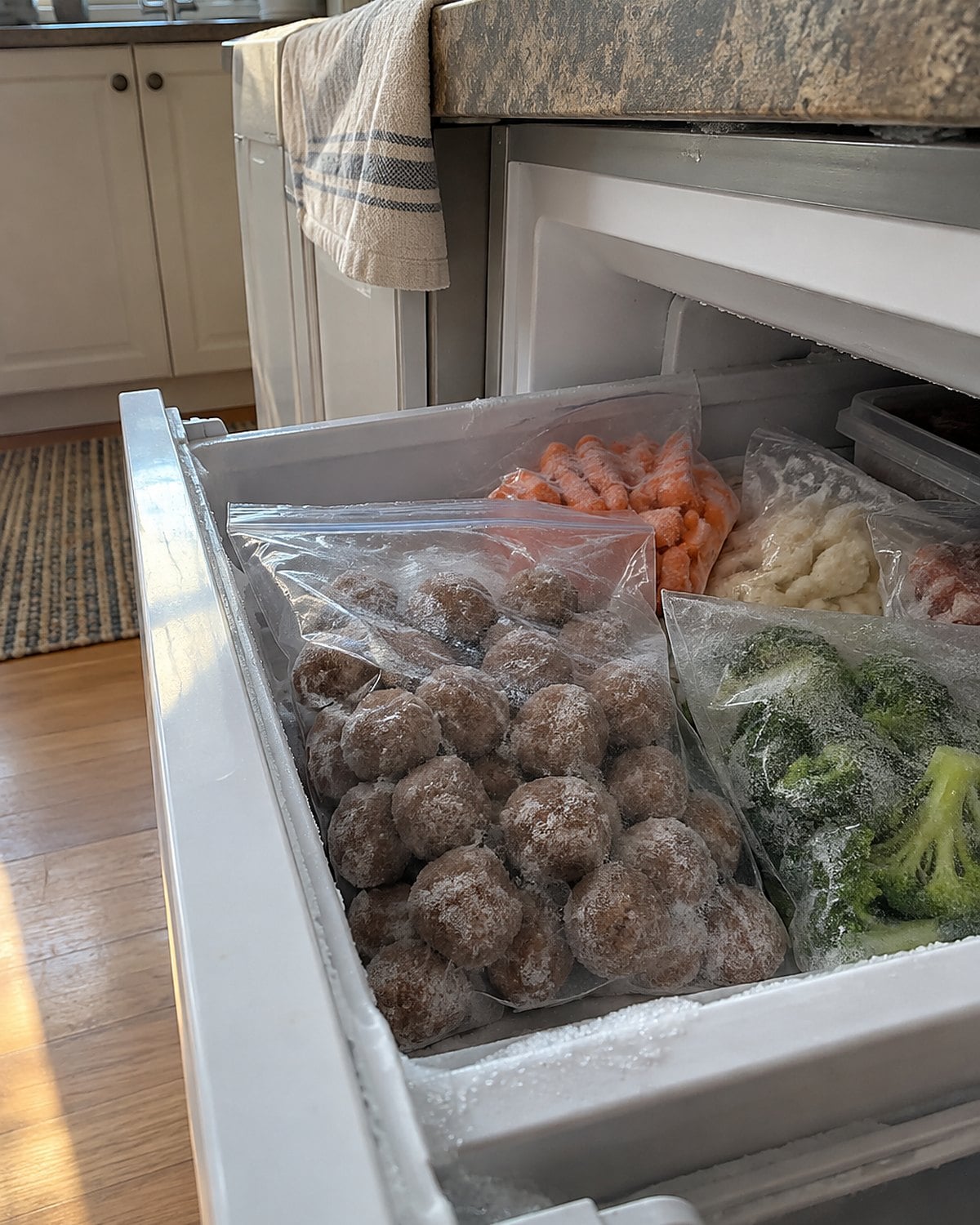 A freezer drawer open to a labeled bag of meatballs beside frost-dusted vegetables.