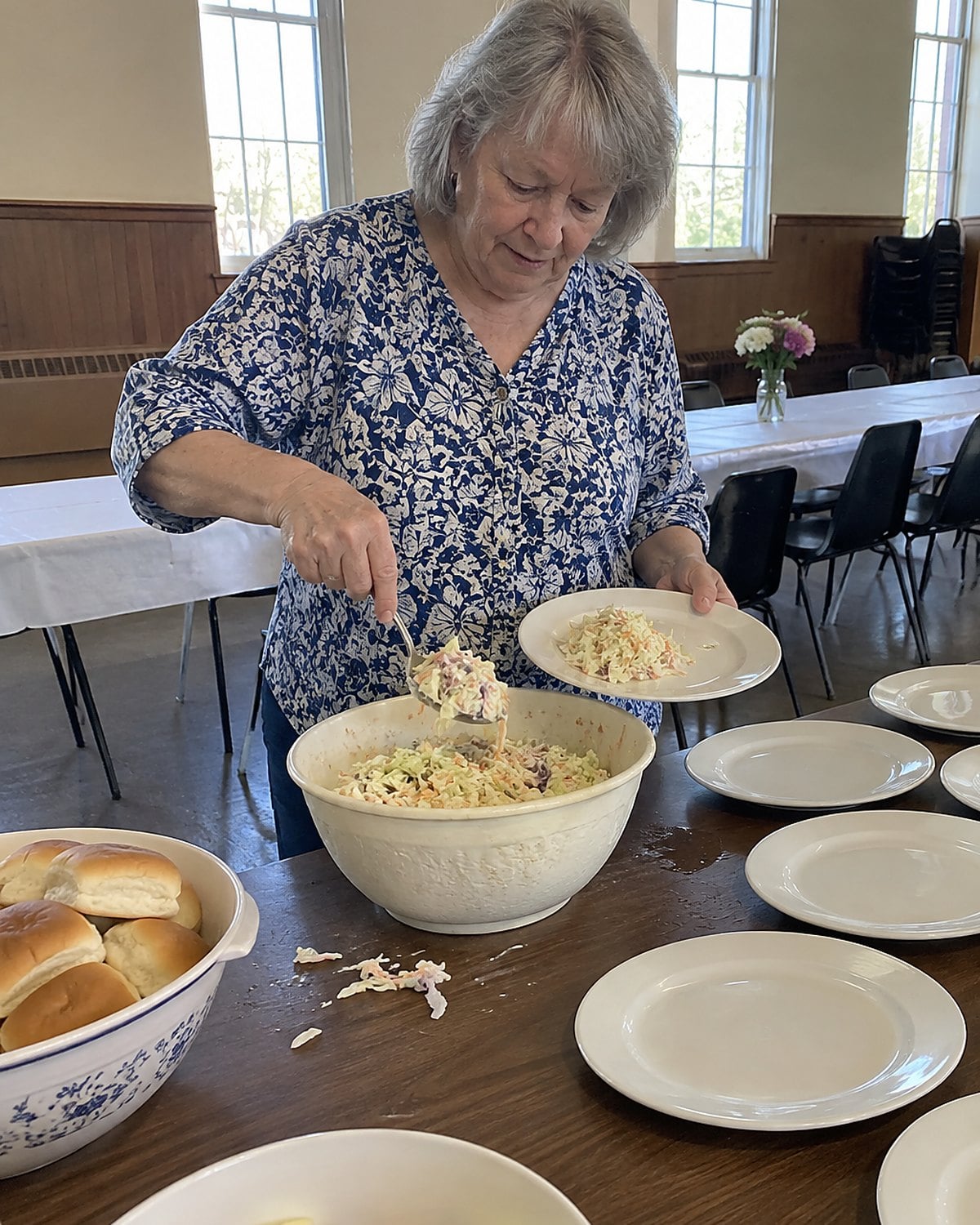 Hands serving coleslaw from a scratched bowl on a church hall table.