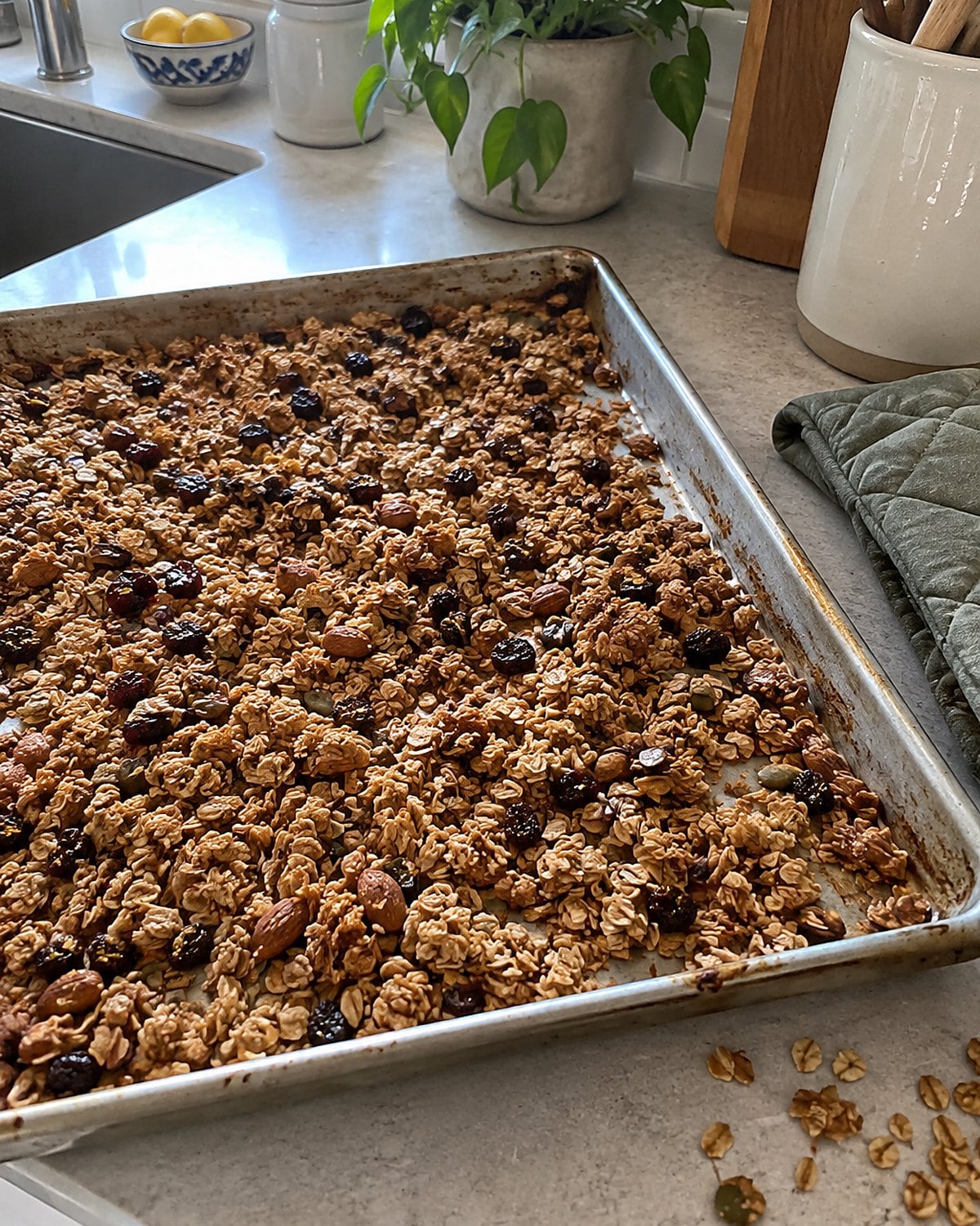 A baking sheet of granola cooling beside an oven mitt and scattered oats.