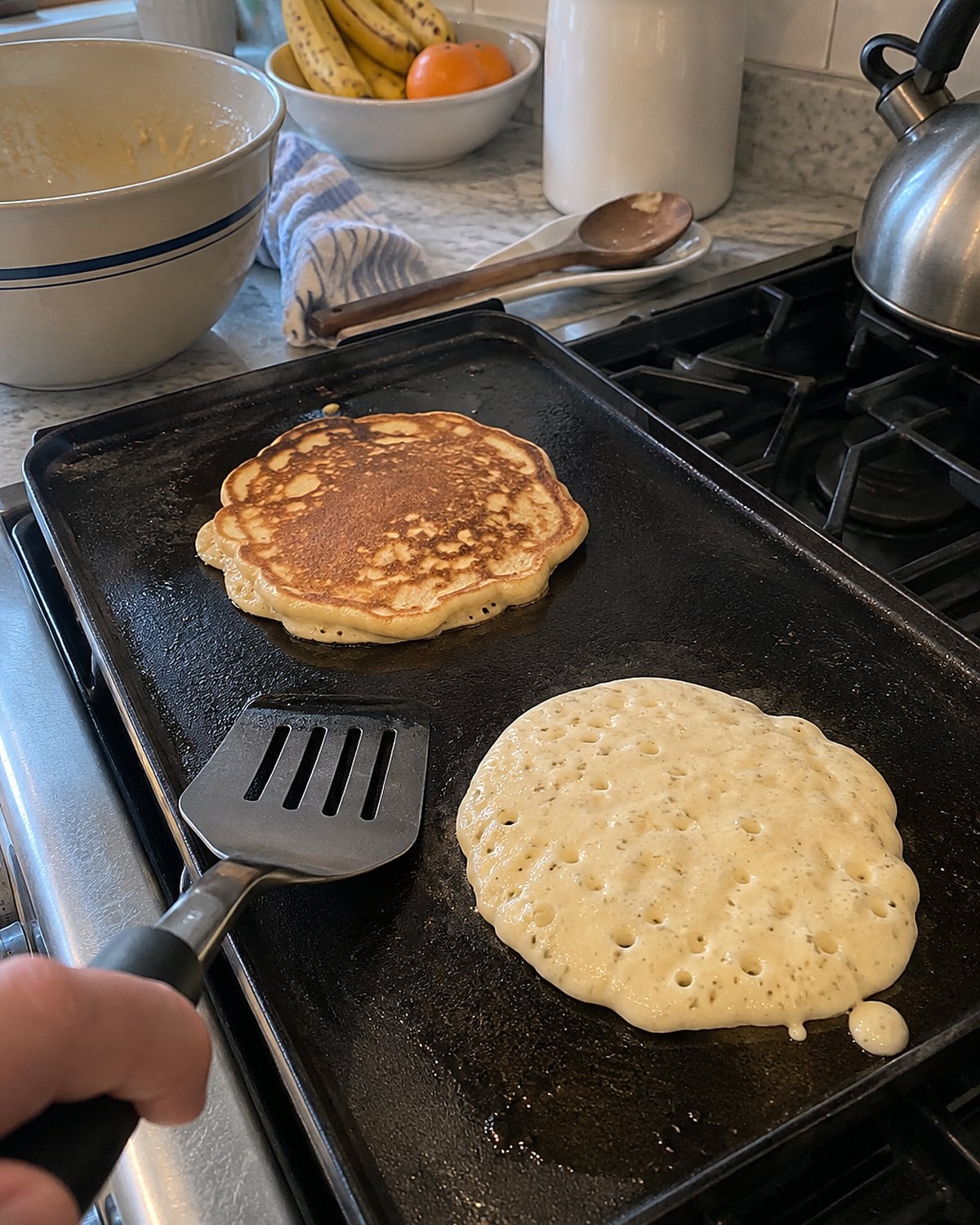 Two uneven pancakes on a griddle, one showing bubbles ready to flip.