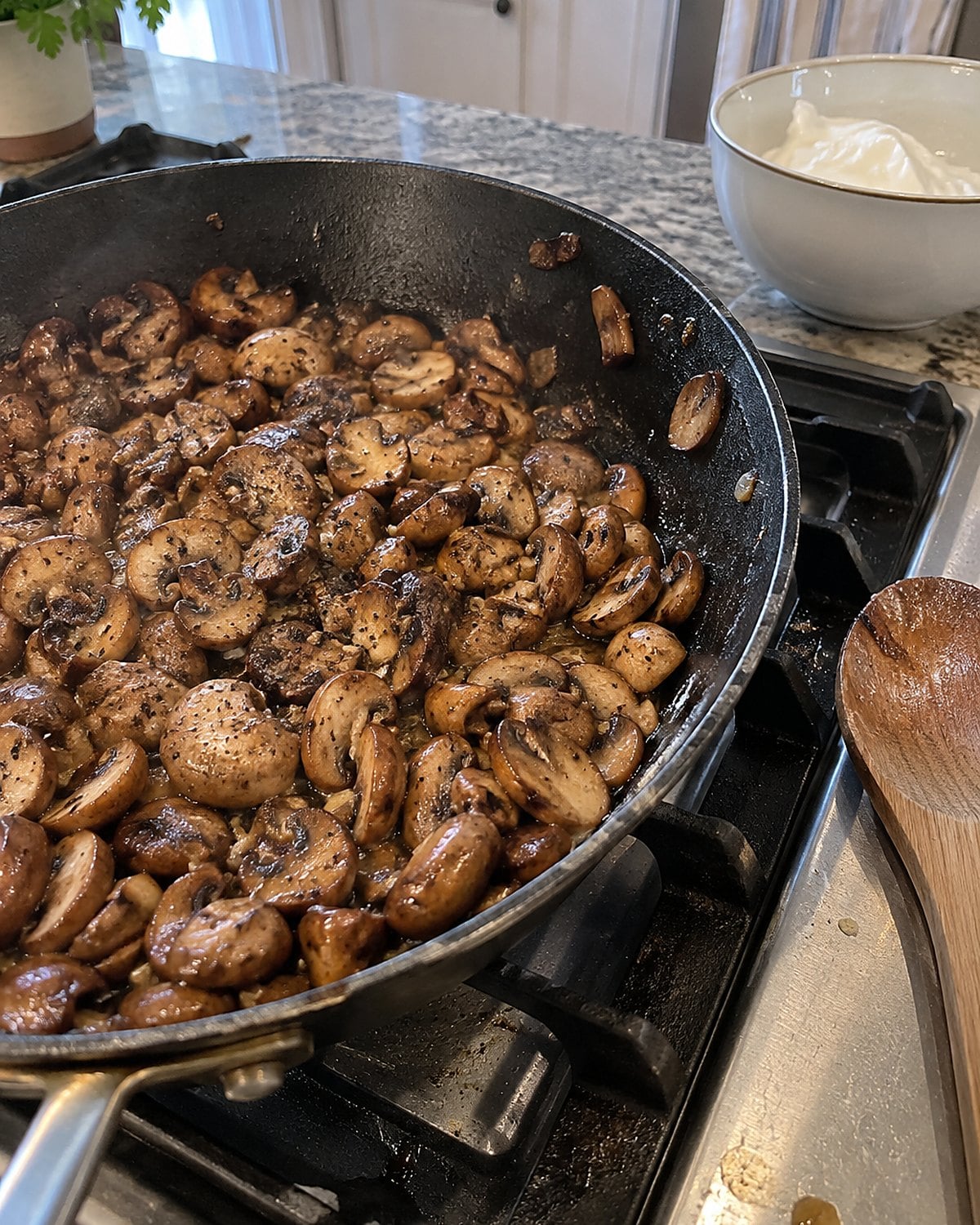 Mushrooms browning in a skillet beside a bowl of sour cream on the stove.