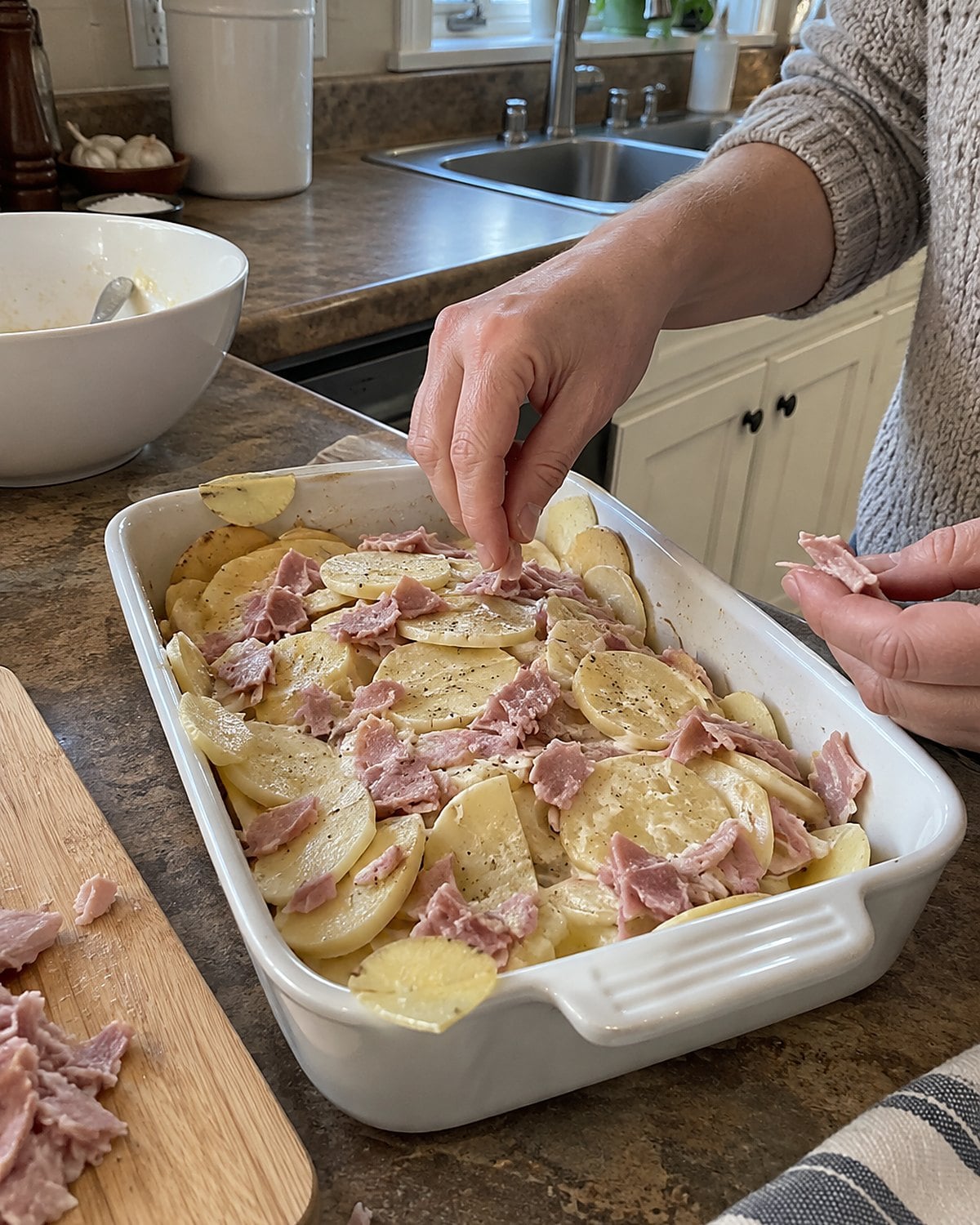 Thin potato slices and scraps of ham layered in a baking dish on the counter.