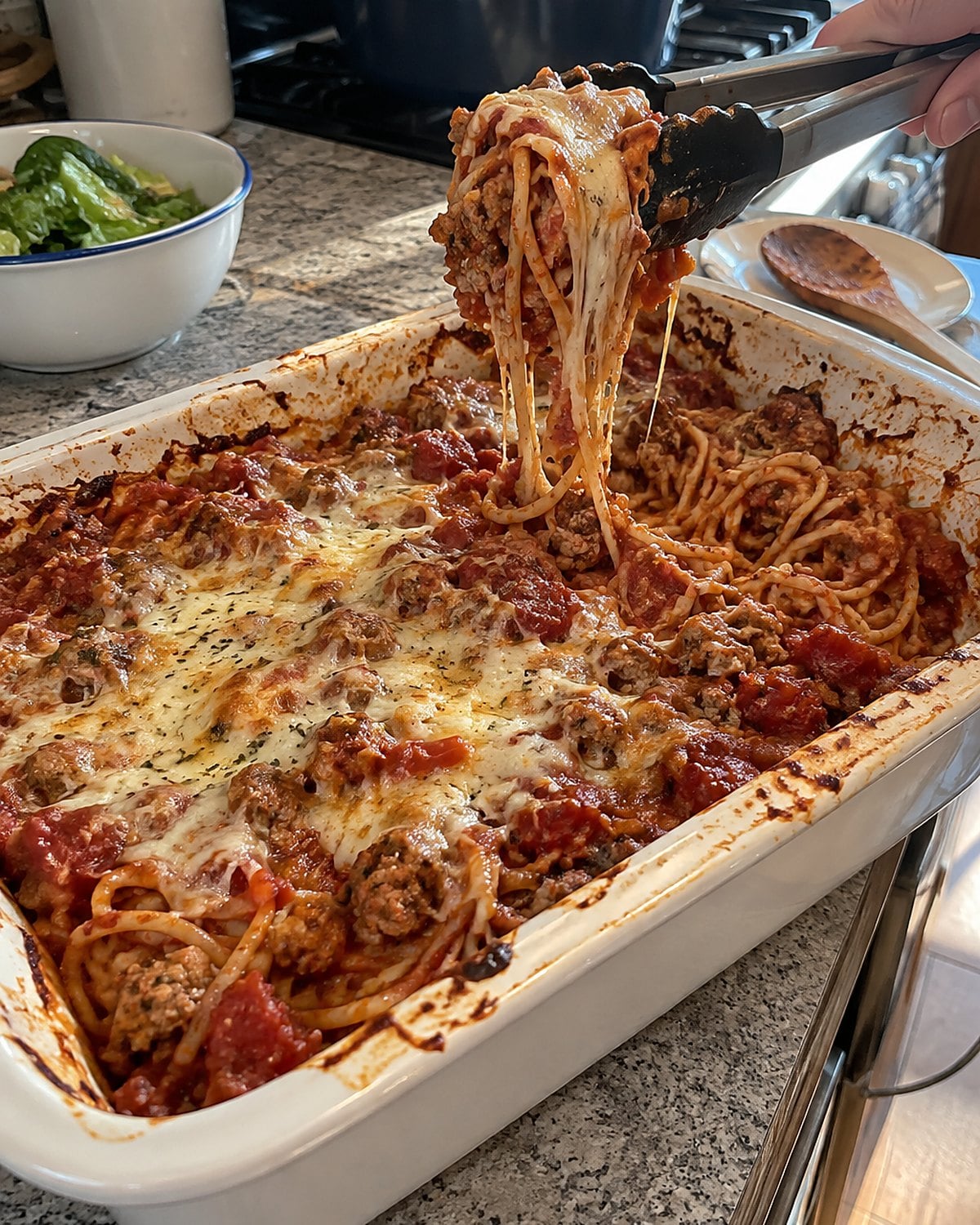 Tongs lifting sauced spaghetti and turkey from a casserole dish beside the stove.
