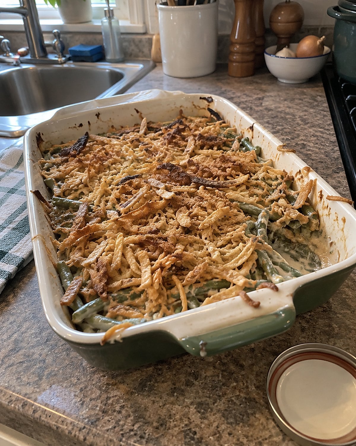 A hand scattering crisp onions over a green bean casserole on a worn kitchen counter.