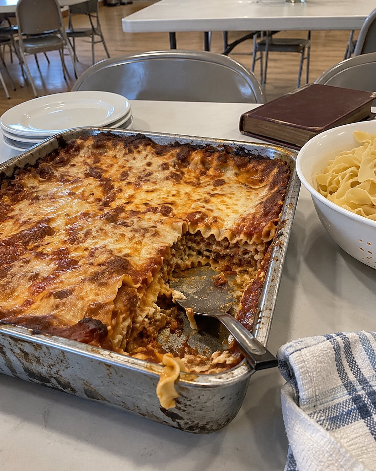 An open, worn church cookbook beside egg noodles and a pan of mock lasagna on a church hall table.