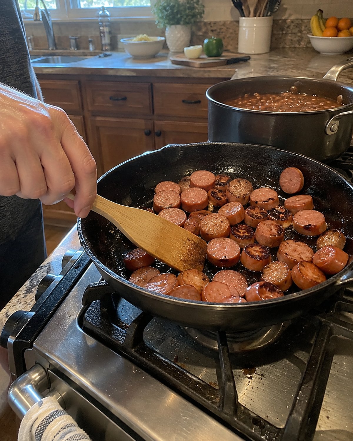 Sliced frankfurters browning in a skillet next to a pot of beans.