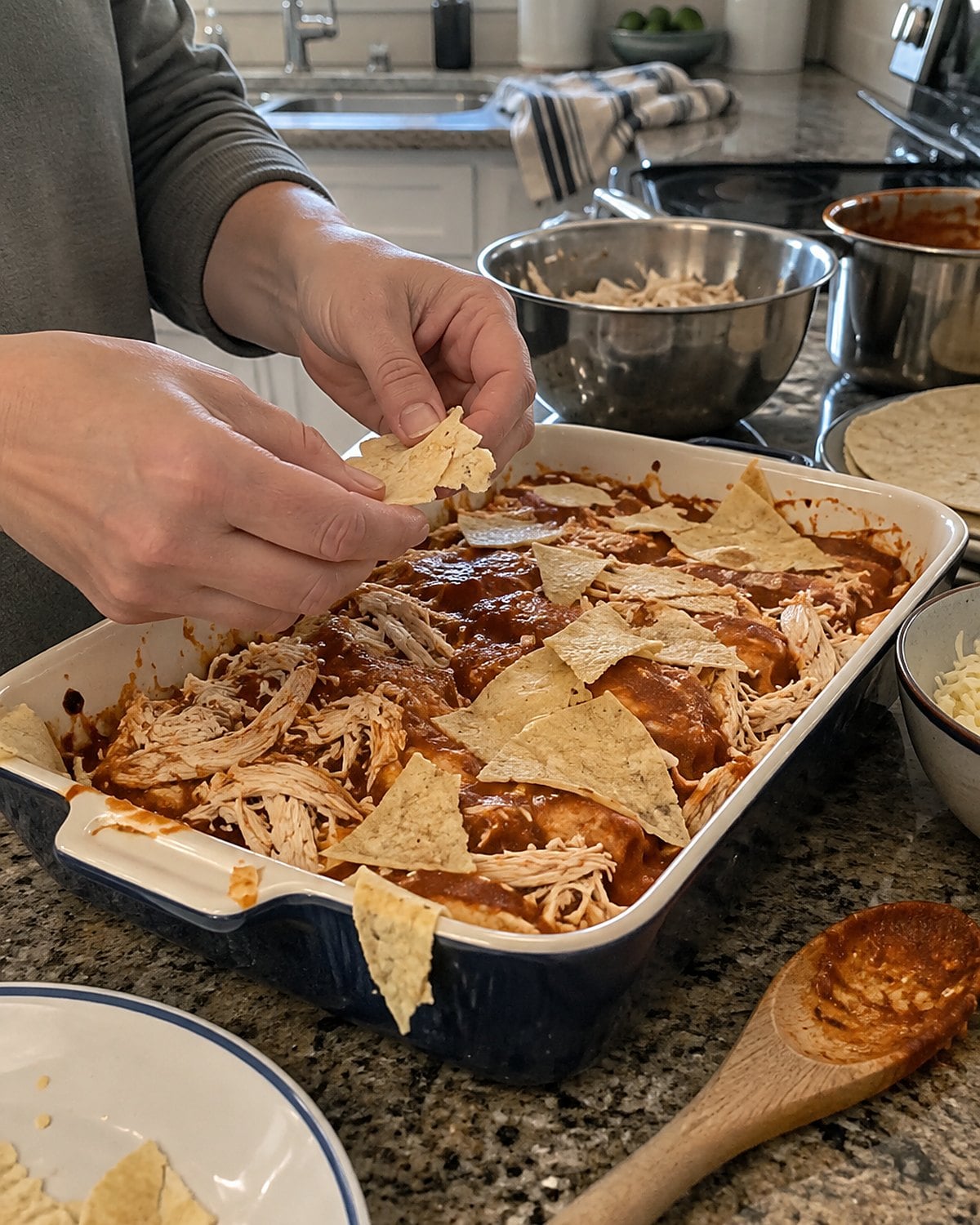 Torn corn tortillas layered with chicken and sauce on a busy kitchen counter.