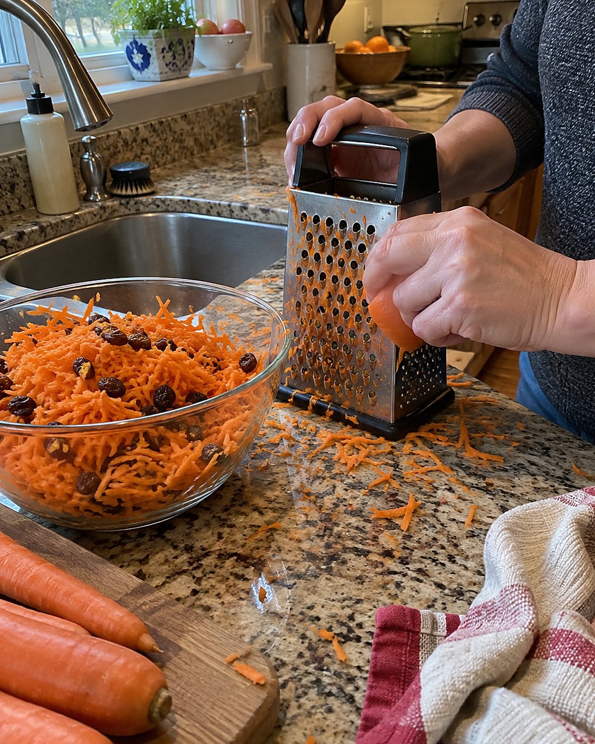 Freshly grated carrots and raisins in a glass dish beside a box grater at the sink.