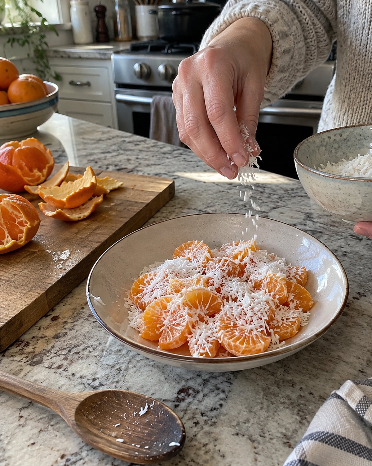 Peeled orange slices under coconut in a shallow dish beside orange peels on a cutting board.