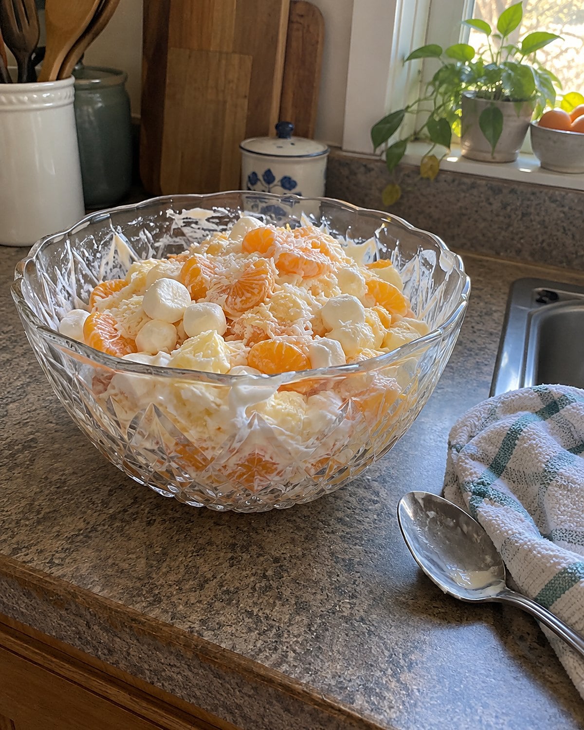 Ambrosia with orange segments in a cut-glass bowl on a kitchen counter, with a spoon resting nearby.