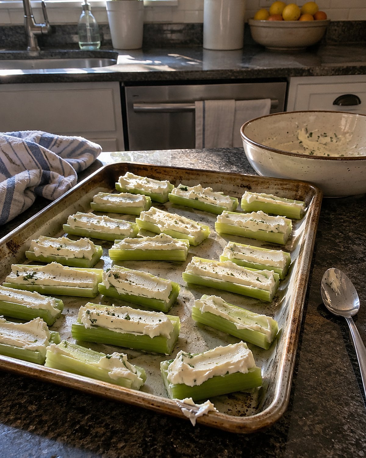 Celery sticks filled with cream cheese on a slightly worn tray beside a teaspoon on the kitchen counter.