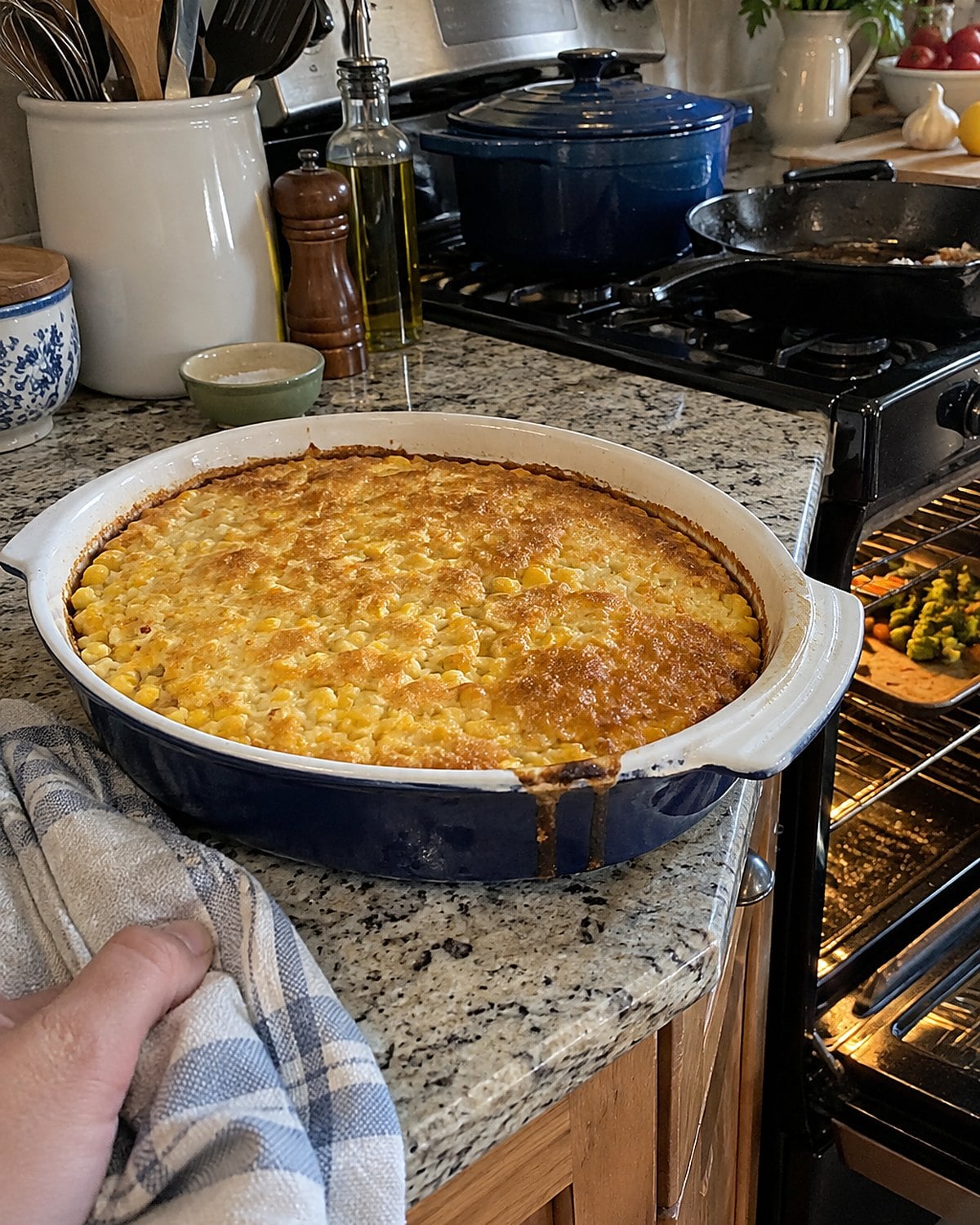 A corn pudding with a browned top waiting on the counter beside a crowded oven.