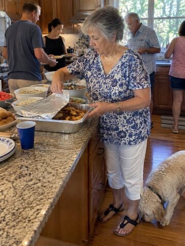 At a picnic table at a family reunion mid-morning, a woman reaches for a foil pan while a dog noses around beneath her feet and relatives pass dishes behind her.